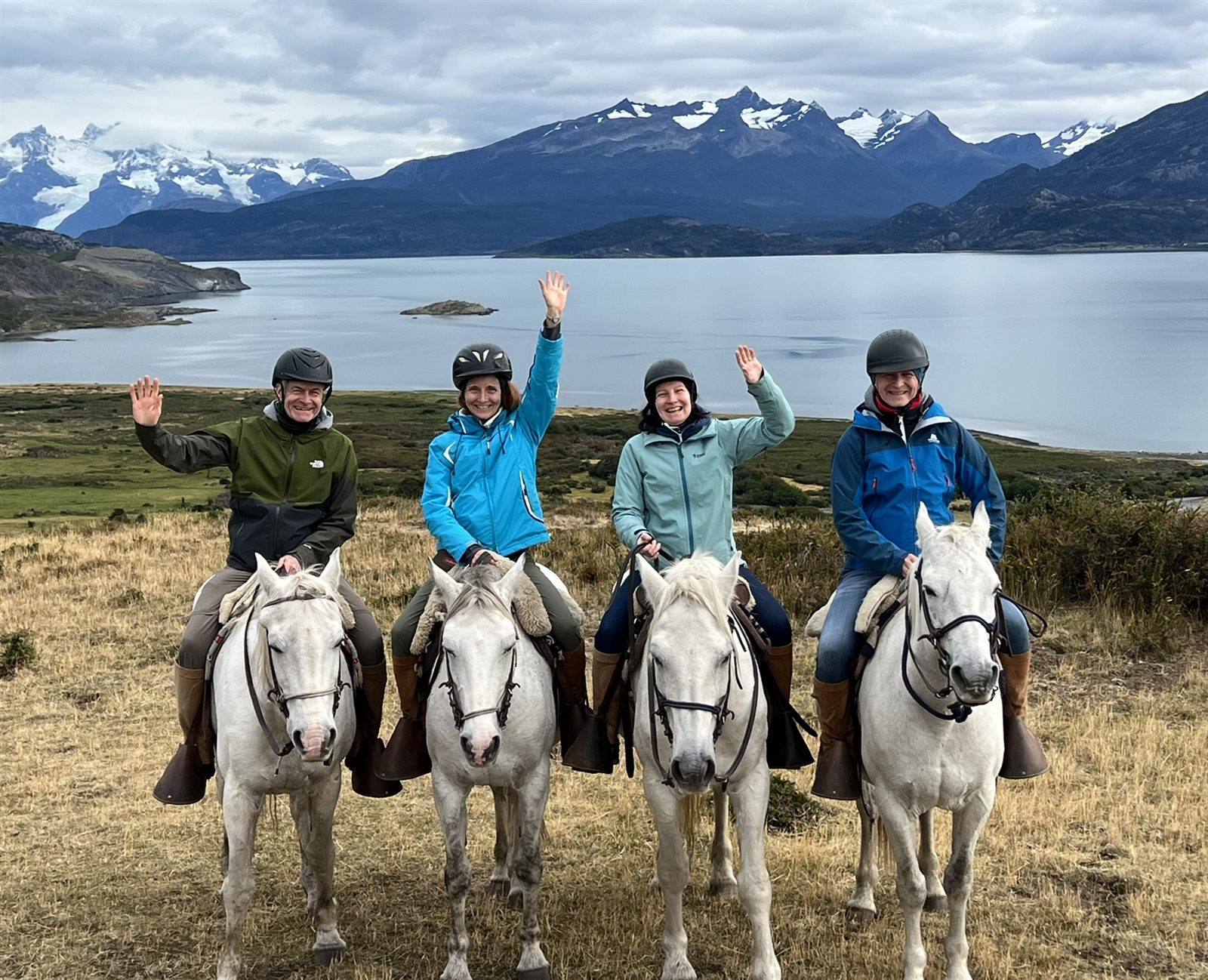 Four riders on white horses waving, with a Patagonian lake and mountains behind them