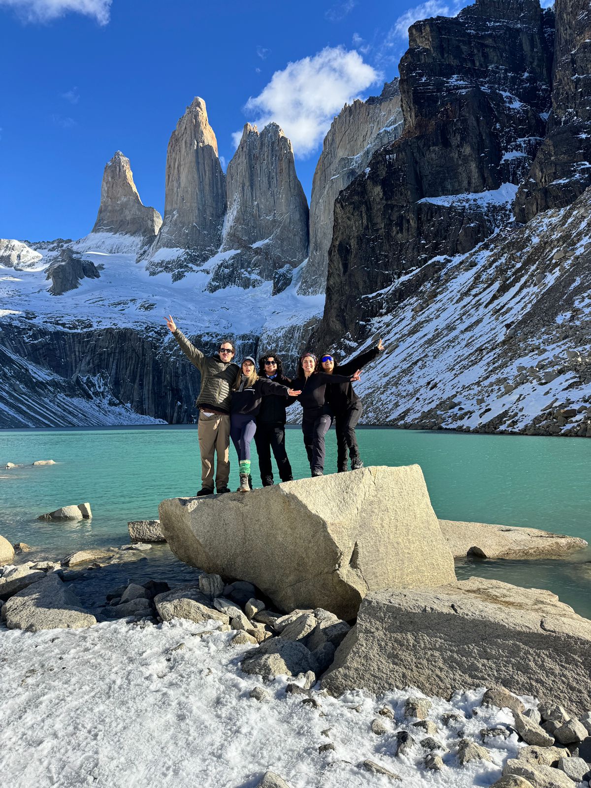 A group celebrating with arms raised on a rock at the turquoise base lake beneath the granite towers of Torres del Paine